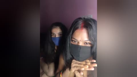 Video of two women with dark skin, long black hair, and masks, standing closely together. One woman in the foreground has a red dot on her forehead. The background is dark and indistinct.