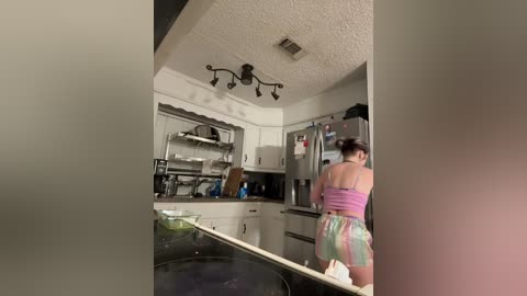 Video of a woman in a pink top and colorful shorts standing at an open kitchen door, with a modern kitchen in the background featuring stainless steel appliances and white cabinets.