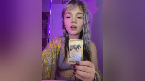 A young, fair-skinned woman with platinum blonde hair, wearing a sheer, yellow floral robe, takes a selfie with a phone, showing a card featuring a tropical beach scene. She has light makeup and red-painted nails. The background is dimly lit with purple lights.