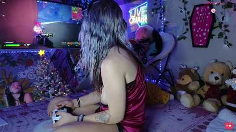 Video of a young woman with long, silver hair, wearing a red satin dress, playing a video game in a dimly lit room filled with plush toys, fairy lights, and a TV screen displaying game footage.