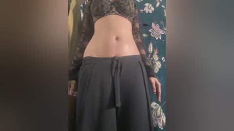 Video of a person with light skin, wearing a black lace crop top and dark gray loose pants, standing against a floral-patterned backdrop.