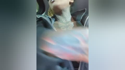 Video of a tattooed person in a car, wearing a black jacket and a grey turtleneck, with blurry, colorful text on their arm, likely a tattoo.