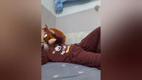 A video of a person in a red and white striped pajama set, lounging on a bed with a teddy bear head prop, in a dimly lit, cozy bedroom with light blue walls.
