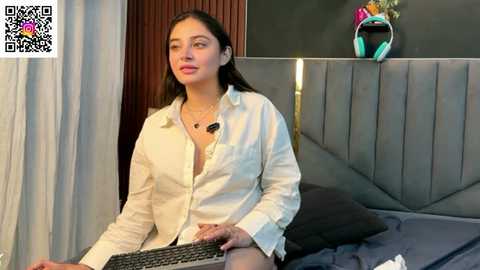 A video of a woman with long dark hair, wearing an unbuttoned white shirt, sitting at a desk with a headset and keyboard, in a modern bedroom with gray headboard and colorful headphones.