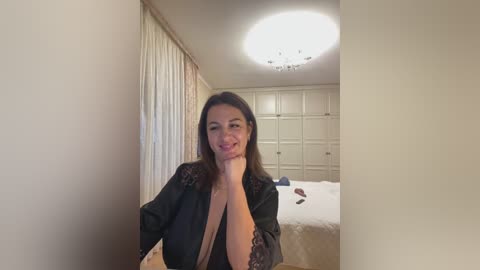 Media: Video of a smiling woman with long dark hair, wearing a black lace robe, sitting in a softly lit bedroom with white walls, curtains, and a bed.