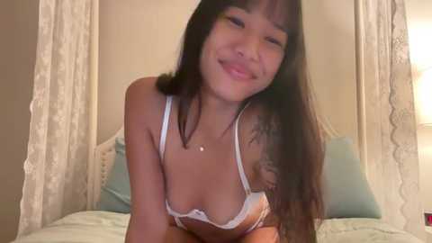 Video of a young Asian woman with long black hair, wearing a white lace bra, smiling in a cozy bedroom with white curtains and light-colored bedding.