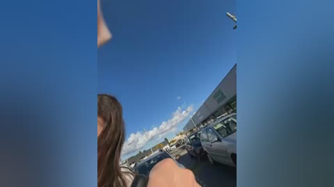 Video of a woman with long brown hair, seen from behind, driving a car on a clear day. She's wearing a white shirt. The sky is blue, and there are parked cars in the background.