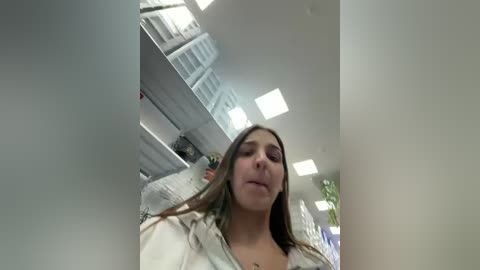 A video of a young woman with straight brown hair, wearing a white sweater, looking down and smiling. She stands in a modern, brightly-lit room with white walls, large windows, and shelves in the background.