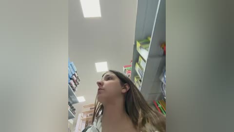 A video of a young Caucasian woman with long brown hair, wearing a white blouse, standing in a well-lit, modern pantry with white walls and shelves filled with various food items and boxes.