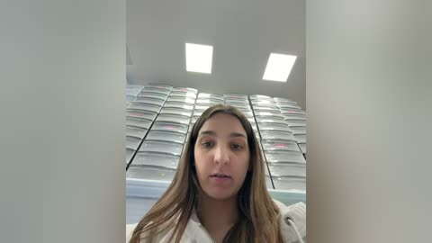 Video of a young Latina woman with long, straight brown hair, wearing a white bathrobe, standing in a modern, white-walled room with large, rectangular, white storage boxes on the left and right walls.