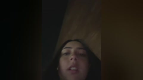 A dimly lit video captures a young woman with fair skin and long dark hair, her face slightly tilted upward, eyes closed, and mouth open as if in a moment of surprise or awe. The background is a dark, blurry wooden surface.