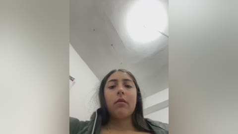 Video of a young Latina woman with long dark hair, medium complexion, wearing a green jacket, looking down at her phone in a white-tiled bathroom with a fluorescent light fixture above.