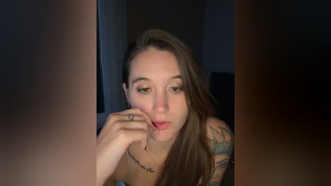 Video of a young woman with long brown hair, fair skin, and tattoos, looking seductively into the camera, wearing a black top, with a dimly lit background.