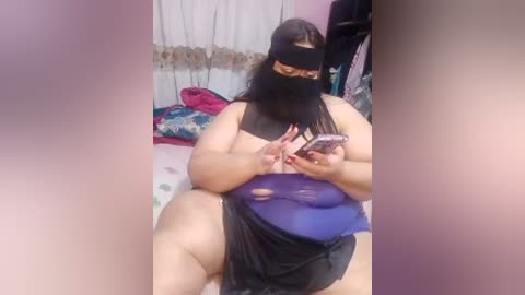 Video of a plus-sized woman with long black hair, wearing a black mask, a blue dress, and black panties, sitting on a bed, texting on a phone.