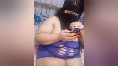 A video of a plus-sized woman with a black blindfold, wearing a revealing blue one-piece swimsuit with cutouts, taking a selfie with a smartphone.