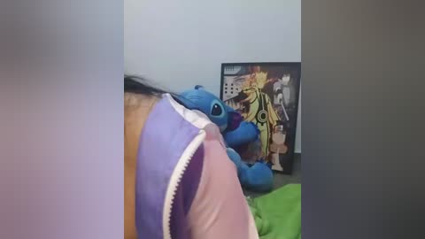 A video of a plush blue alien toy lying on a bed with a green sheet, next to a framed anime character poster.