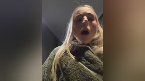 Video of a blonde woman with light skin, wearing a textured green sweater, her mouth open in apparent surprise or shock, set in a dimly lit, possibly indoor, space.