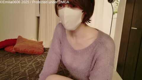 Video of a woman with short brown hair, wearing a white face mask and a light purple sweater, sitting on a patterned couch in a dimly lit room.
