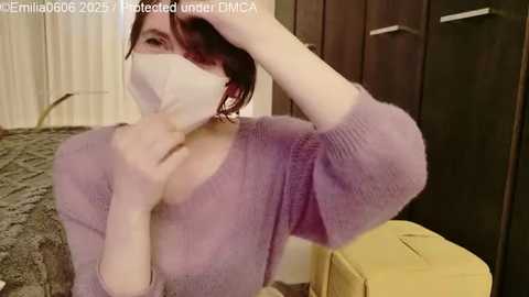 A video of a woman with short red hair, wearing a purple sweater, a face mask, and a yellow bag, seated on a patterned carpet. She covers her mouth with one hand.