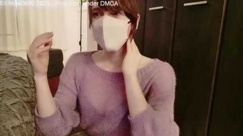 Video of a young woman with short brown hair, wearing a lavender knit sweater, face mask, and black phone, seated indoors.