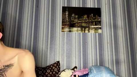 Video of a man with a tattooed back, standing in a room with striped wallpaper and a framed cityscape photo. He is facing away, partially visible.