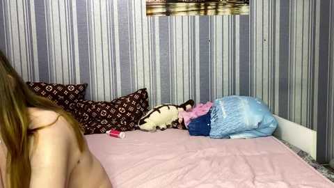 Video of a bedroom with striped wallpaper, a pink bedspread, and a woman's back, showing bare shoulders. Pillows with geometric patterns and a blue blanket are visible.