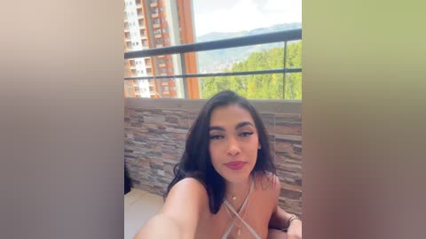 Video of a young woman with long dark hair and fair skin, wearing a white bralette, sitting on a balcony with a stone wall background and cityscape in the background.