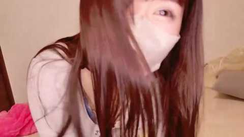 Video of an Asian woman with long, straight black hair, wearing a white mask and a light-colored jacket, leaning forward in a dimly lit room with beige walls and a pink blanket.