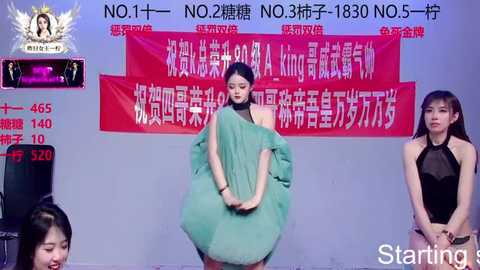A video captures three East Asian women in a runway setting. One models a voluminous, green, feathered gown, another wears a black halter dress, and a third stands to the side. Red banners with Chinese text are visible in the background.