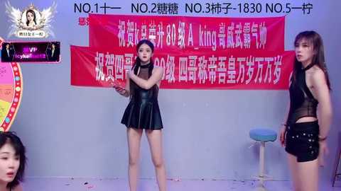Video of three East Asian women in provocative black outfits, standing in front of a red banner with Chinese text. The setting is indoors, with a white floor and a stool.