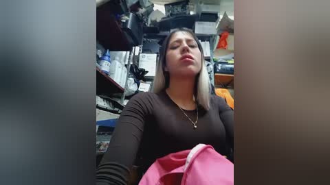Video of a young Latina woman with medium skin tone and straight black hair, wearing a black top, seated in a cluttered, dimly-lit room with shelves filled with various items.