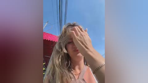 A video of a woman with long blonde hair, wearing a sleeveless top, shielding her eyes with her left hand, standing in front of a red-roofed building against a clear blue sky.