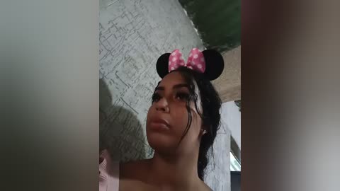 Media: Video of a young Black woman with medium brown skin and short curly hair, wearing a pink polka-dot bow headband and black Minnie Mouse ears, in a dimly lit room with gray brick walls.