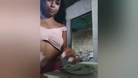 Media: Video of a dark-skinned woman with curly hair, wearing a pink off-shoulder top, washing dishes in a rustic kitchen with a tin roof and concrete walls.