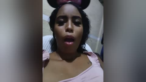 Video of a young Black woman with medium-dark skin, wearing a light pink off-shoulder top, her mouth slightly open, and a headband. The background is blurred.