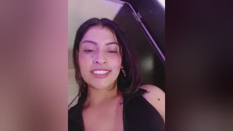 Video of a smiling woman with medium-dark skin tone, medium-length black hair, and hoop earrings, wearing a black sleeveless top, taken indoors with dim lighting.