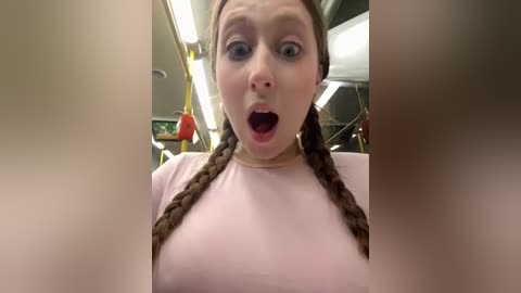 A video of a young woman with light skin and brown pigtails in a pink shirt, wide-eyed and open-mouthed, seated in a subway car.