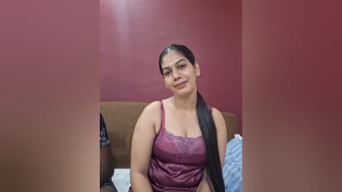 Video of a smiling Latina woman with medium-brown skin and long black hair, wearing a burgundy lace camisole, seated on a brown couch against a maroon wall.