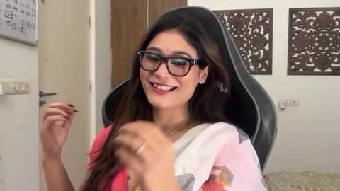 Video of a smiling South Asian woman with long black hair, wearing glasses, a pink sari, and black headband, seated in a black chair in a modern, white room with wooden furniture and decorative art.