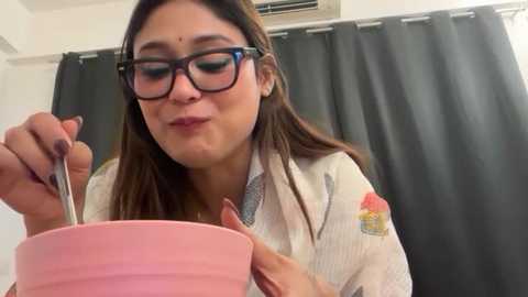 Video of a smiling Latina woman with long brown hair, glasses, and a white blouse with colorful embroidery, eating from a pink bowl using a spoon. She's indoors near a dark curtain and an air conditioning unit.