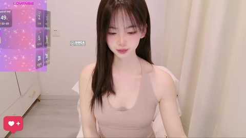 A video of an Asian woman with straight black hair, fair skin, and small breasts, wearing a beige tank top, sitting indoors. The background features a white curtain and a purple digital display.