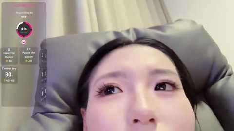 Video of an Asian woman with straight black hair, lying on a gray pillow, eyes closed, in a cozy room with beige curtains.