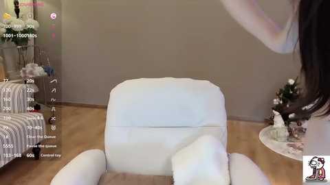 A video captures a woman in a white, plush armchair in a minimalist living room. The room features a beige wall, a floral-patterned sofa, and a small table with decorative items. The image includes a \"Chat\" overlay with a virtual chatbot's face.