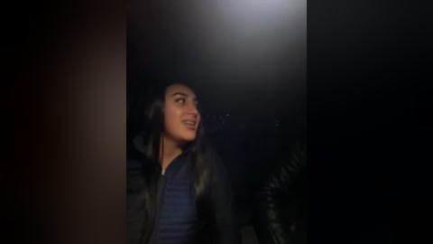 Video of a woman with long, dark hair and a dark jacket, smiling, standing in a dimly lit outdoor setting with trees in the background.