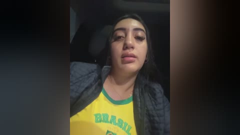 Video of a Latina woman with medium brown skin and long dark hair, wearing a yellow T-shirt with green text and a gray plaid shirt, sitting in a dimly lit room.