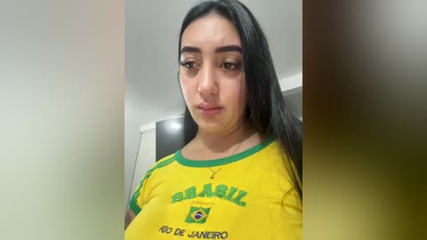 Video of a young woman with long black hair, wearing a yellow Brazil soccer jersey, taken indoors, blurred background.