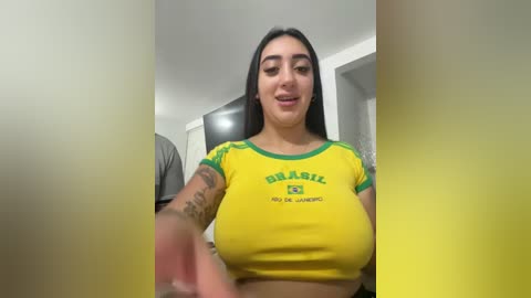 A video of a young woman with long black hair, wearing a tight yellow crop top with green accents and a Brazilian flag, smiling, in a modern living room.