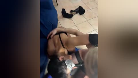 Video of a man lying on a blue couch with a woman on top, her breasts exposed, wearing a black bra, and a man filming them.