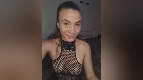 A video of a smiling, fair-skinned woman with long brown hair tied back, wearing a black fishnet top that reveals her breasts. The background is dimly lit, featuring a dark, patterned couch.
