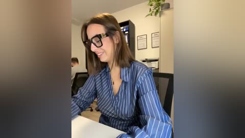 A video of a Caucasian woman with shoulder-length brown hair, glasses, and a blue pinstriped blouse, smiling, sitting at a desk in a modern office.